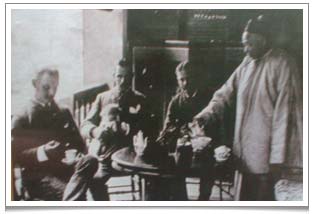 Western officials enjoying a cup of tea at a Chinese teahouse.  Note that the gentleman on the left is holding a Western cup and saucer whereas on the table on the right there is a Chinese gaiwan tea cup.